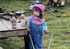 A small toddler in a pink hat and overalls is drinking a juice box outside.