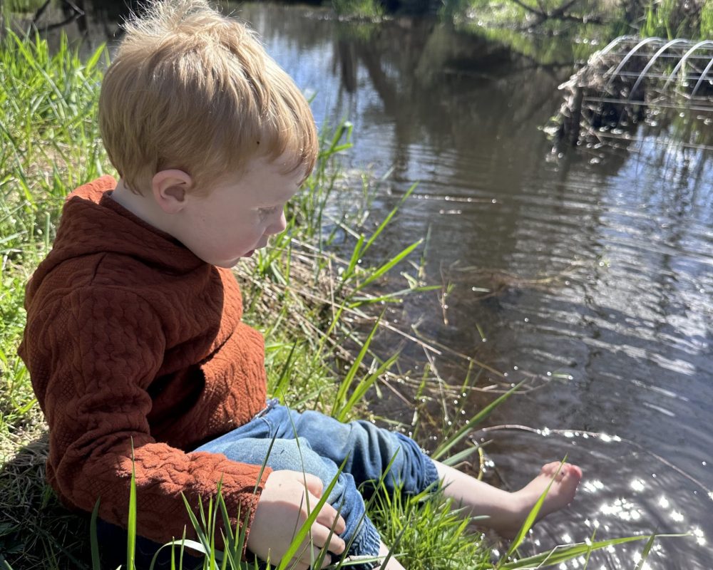 A young boy sits near a riverbank in his bare feet.